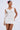 A woman poses in the NEVER BASIC RUFFLED MESH MINI DRESS, a sleeveless, white bodycon mini dress with textured ruffled details. She wears a pink and purple flower in her hair and stands against a plain light background.