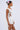 A woman in profile wears the NEVER BASIC RUFFLED MESH MINI DRESS, featuring a textured bodycon silhouette with ruffled mesh and draping. She styles her hair in a bun with pink and purple flowers and wears large gold hoop earrings.