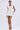 A woman faces forward in the NEVER BASIC RUFFLED MESH MINI DRESS—short, fitted, and white with textured mesh and ribbon shoulder details. She wears a flower in her hair, hoop earrings, beige platform heels, all set against a plain light gray background.