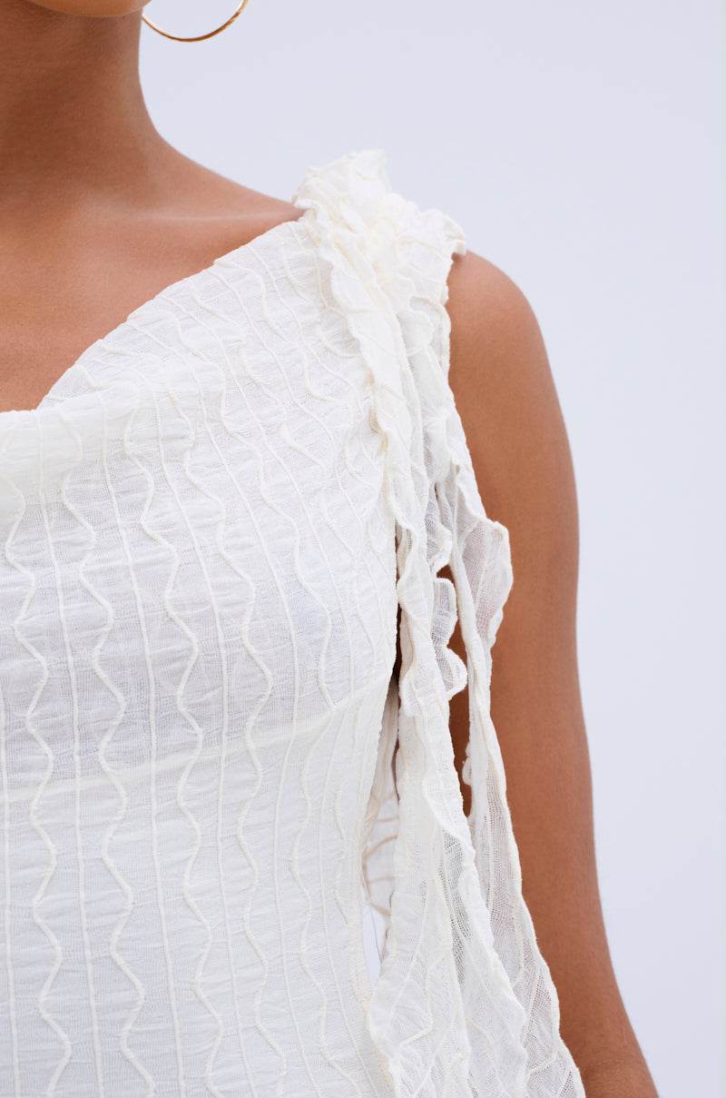 A close-up of a person with medium brown skin wearing the NEVER BASIC RUFFLED MESH MINI DRESS, a white textured, sleeveless dress with ruffled detail on one strap. They accessorize with a large gold hoop earring against a light blue background.