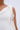 A close-up of a person with medium brown skin wearing the NEVER BASIC RUFFLED MESH MINI DRESS, a white textured, sleeveless dress with ruffled detail on one strap. They accessorize with a large gold hoop earring against a light blue background.