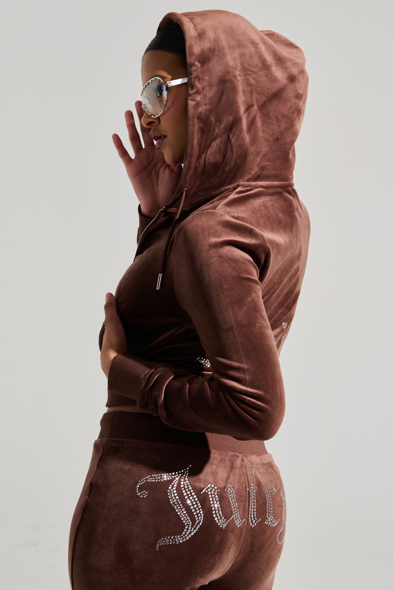 A person wearing the SOLID CLASSIC JUICY HOODIE WITH BACK BLING and matching pants, with "Juicy" emblazoned in rhinestones on the back. They sport sunglasses with their hood up, captured in a profile view against a plain background. This Y2K-inspired outfit channels the iconic Juicy Couture Hoodie era perfectly. 2