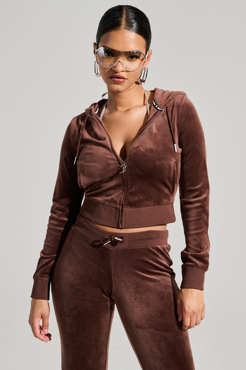 A woman with her hair tied back, adorned with large hoop earrings and chic sunglasses, strikes a confident pose in a Y2K-inspired brown velour tracksuit that brings to mind the SOLID CLASSIC JUICY HOODIE WITH BACK BLING. The backdrop is simple and light-colored. 3