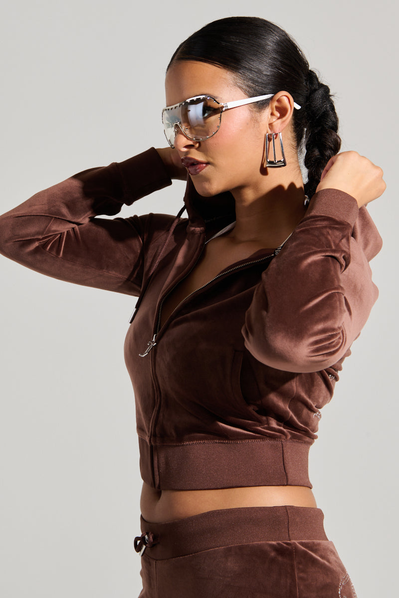 A person wearing a dark brown SOLID CLASSIC JUICY HOODIE WITH BACK BLING and matching pants poses with their hands adjusting the hood. Their dark, pulled-back hair complements reflective sunglasses and geometric earrings. The Y2K-inspired look is set against a plain gray background. 4