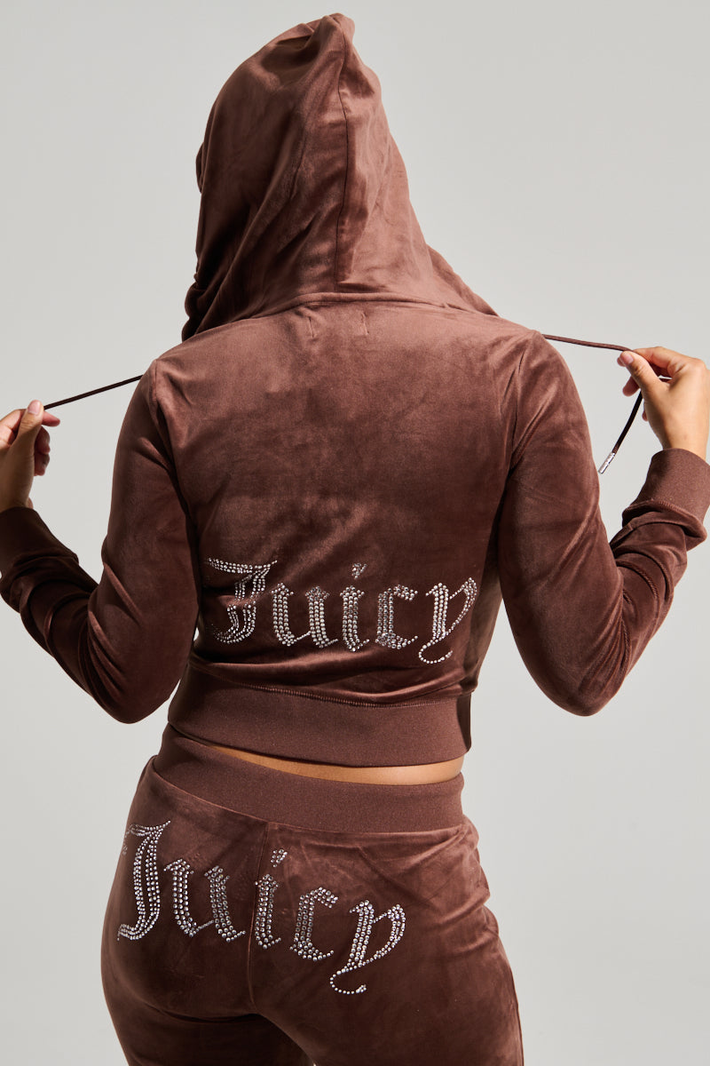 A person facing away from the camera is wearing a Y2K-inspired brown velour tracksuit, featuring the SOLID CLASSIC JUICY HOODIE WITH BACK BLING. The back of the Juicy Couture hoodie and the rear of the pants both showcase "Juicy" adorned in rhinestones. The individual is holding the hoodie strings with both hands. 0