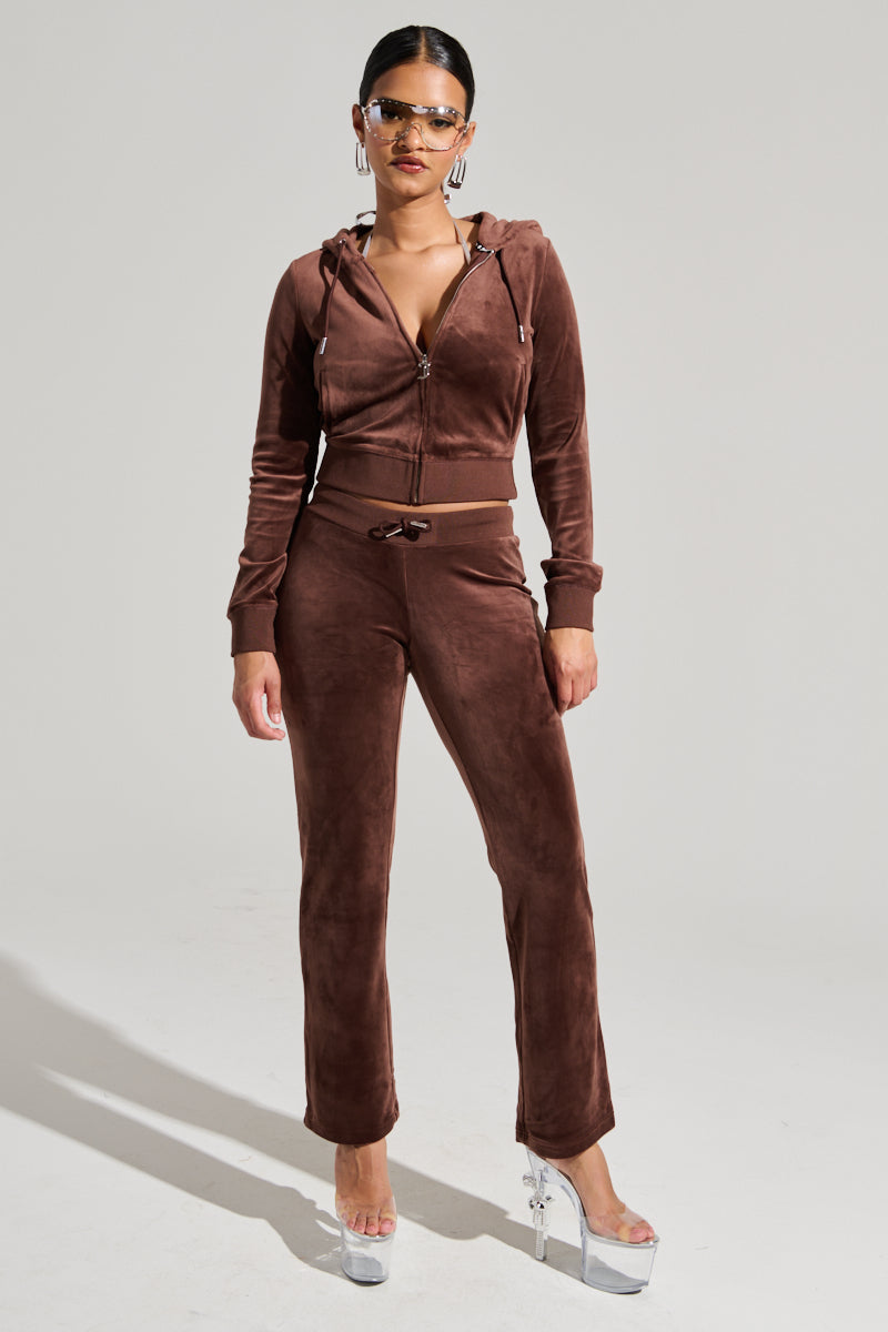A woman dressed in a brown two-piece velour outfit, featuring the SOLID CLASSIC JUICY HOODIE WITH BACK BLING and matching drawstring pants. Her look is completed with large hoop earrings, sunglasses, and shiny platform heels, capturing a Y2K-inspired style. She poses against a simple gray background. 6