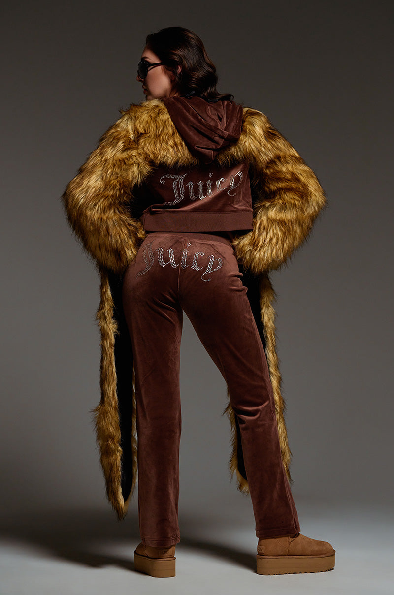 A person wearing a brown Juicy Couture SOLID RIB WAIST VELOUR PANT tracksuit with "Juicy" in rhinestones on the back of the hoodie and pants. The Y2K-inspired outfit showcases a faux fur-trimmed hood and sleeves, accessorized with dark sunglasses and brown platform shoes.