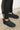 Standing on a concrete surface near a white wall, the individual wears the UGG Ultra Mini Bootie in Black, featuring black suede and a cozy wool lining. These boots are elegantly paired with black skinny jeans.