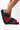 A close-up of a person wearing the UGG WOMENS FLUFF YEAH SLIDE BLACK MULTI slippers. These colorful, fluffy slippers feature a black, red, and pink striped design. The back strap is black and white with the "UGG" logo printed on it. The person's toenails are painted red.