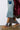 A person in a denim skirt, gray socks, and the UGG TAZZ Flat Slipper in Chestnut walks on a concrete floor. The suede upper of the slippers adds a stylish touch, complemented by their platform sole. They wear a long maroon coat, and the photo focuses on the lower half.