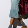 A person in a denim skirt, gray socks, and the UGG TAZZ Flat Slipper in Chestnut walks on a concrete floor. The suede upper of the slippers adds a stylish touch, complemented by their platform sole. They wear a long maroon coat, and the photo focuses on the lower half.