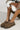 A person wearing hickory UGG Tazz slip-on shoes with a thick sole and patterned trim sits cross-legged on a light surface. They are dressed in a cozy ensemble, featuring a green coat with white fur cuffs.