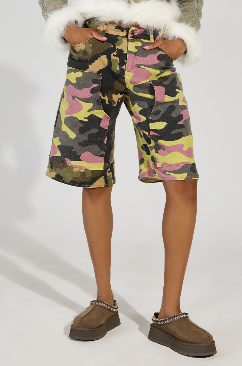 A person is wearing camo-patterned shorts in green, pink, and yellow hues with their hands tucked into the pockets. They are also wearing fuzzy gray sleeves paired with brown UGG Tazz clogs in Hickory featuring decorative stitching, creating a cozy ensemble against a white background.