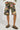 A person is wearing camo-patterned shorts in green, pink, and yellow hues with their hands tucked into the pockets. They are also wearing fuzzy gray sleeves paired with brown UGG Tazz clogs in Hickory featuring decorative stitching, creating a cozy ensemble against a white background.