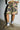 A person wearing pink and green camouflage shorts with hands in pockets, a white fur-trimmed jacket, wool blend socks featuring blue stripes, and brown UGG Tazz in Hickory slip-on shoes. They are standing on a concrete floor against a white brick wall in this cozy ensemble.