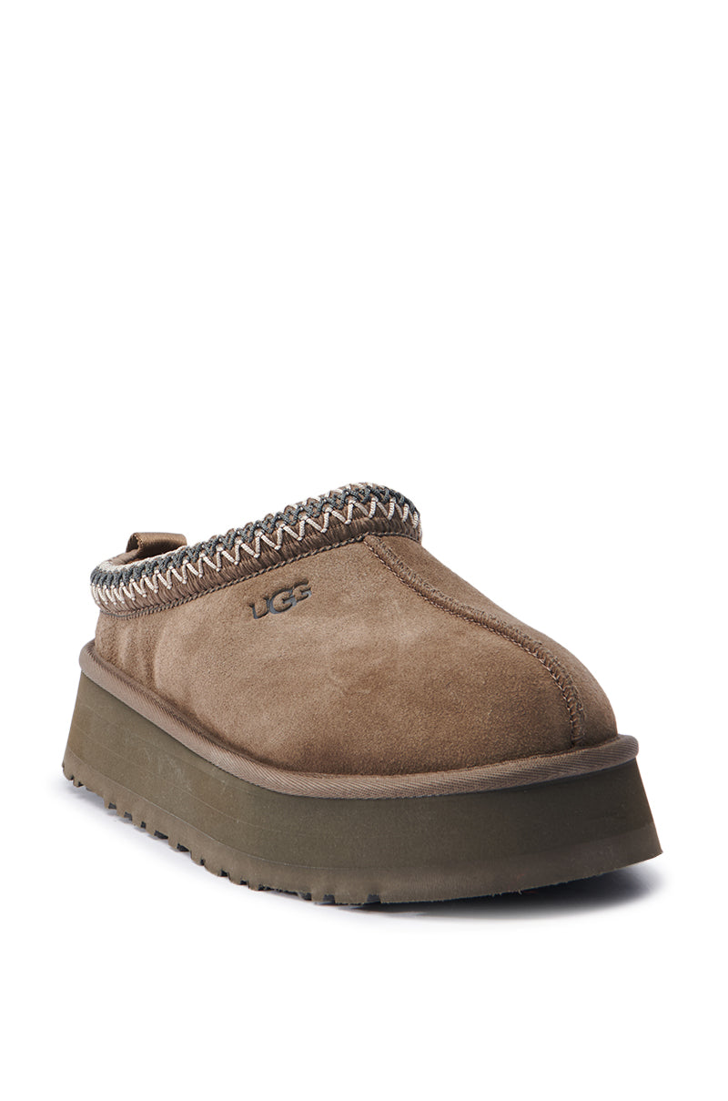 Introducing the UGG Tazz in Hickory: a suede slip-on shoe in a warm brown hue, featuring a thick sole and decorative stitching around the opening. The cozy design is accented with the UGG logo on the side, providing both style and comfort.