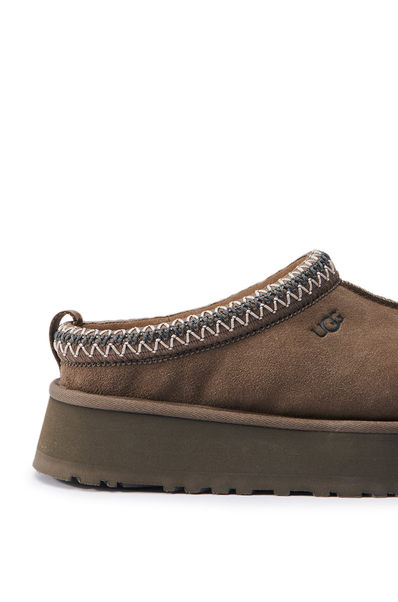 The UGG TAZZ IN HICKORY is a casual slipper with a soft suede exterior and visible stitching around the opening. It features a wool blend for enhanced comfort, a thick flat sole, and a small fabric pull tab at the heel. The UGG logo is embroidered on the side, making it perfect for any cozy ensemble.