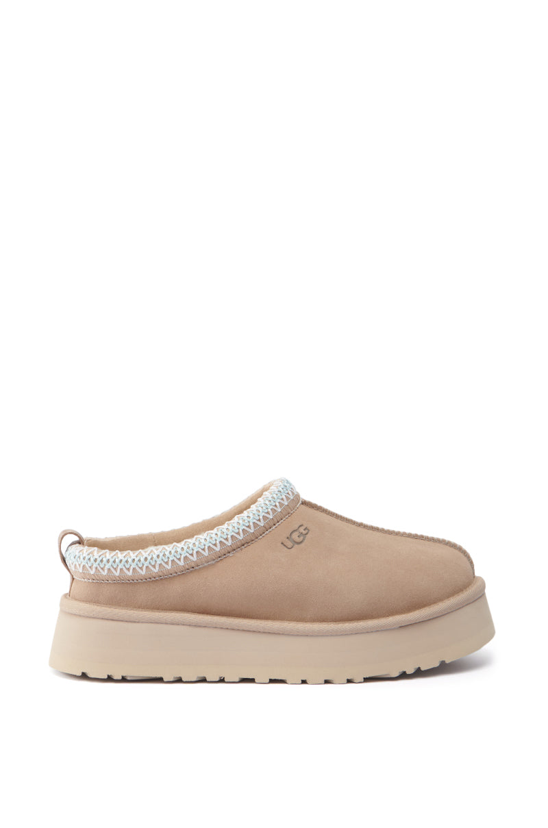 The UGG Tazz Flat Slipper in Brown boasts a tan suede upper complemented by a delightful woven trim along the top edge. It features a convenient pull tab at the back and showcases the iconic UGG logo embossed on the side, offering both style and simplicity with every step.