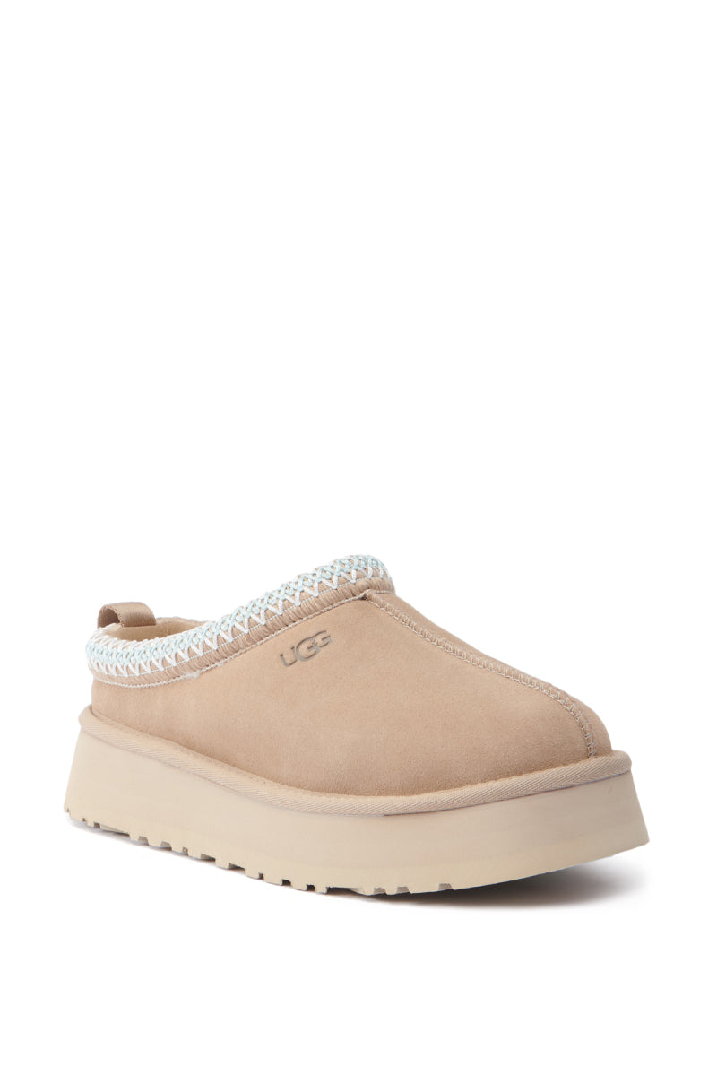 The UGG Tazz Flat Slipper in brown is a single slip-on shoe featuring a suede upper with a platform sole. It includes a white braided detail around the opening and prominently displays the UGG logo on the side, providing comfort with its plush, suede-like texture.