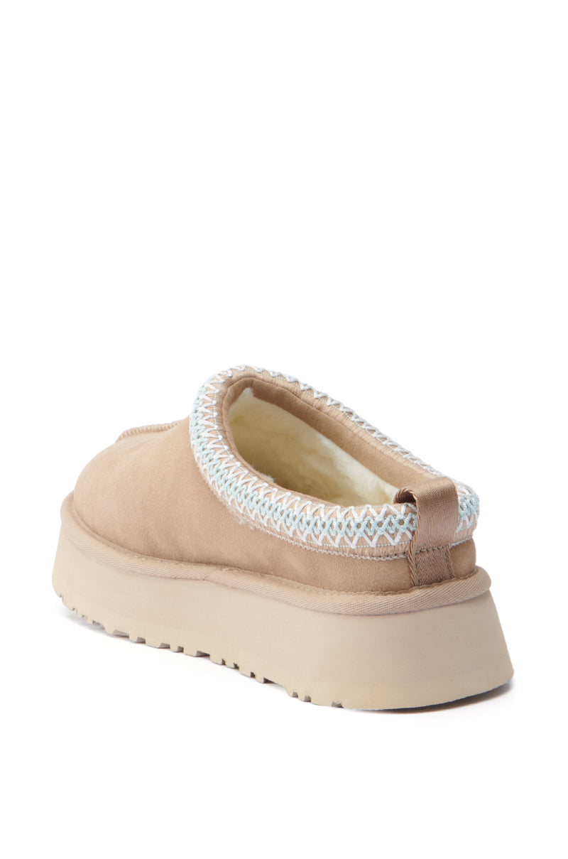 The UGG Tazz Flat Slipper in Brown is a tan suede slip-on shoe with a thick beige platform sole, accented by blue and white stitching around the opening. It features a warm, textured lining and includes a convenient pull tab at the heel for easy wear.