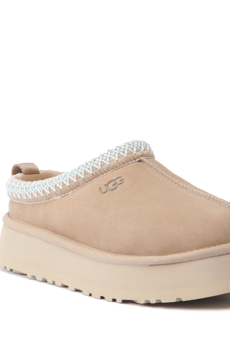 The UGG Tazz Flat Slipper in Brown is a single tan slip-on shoe featuring a suede upper and a woven trim around the opening, complemented by a platform sole and the "UGG" logo on the side.