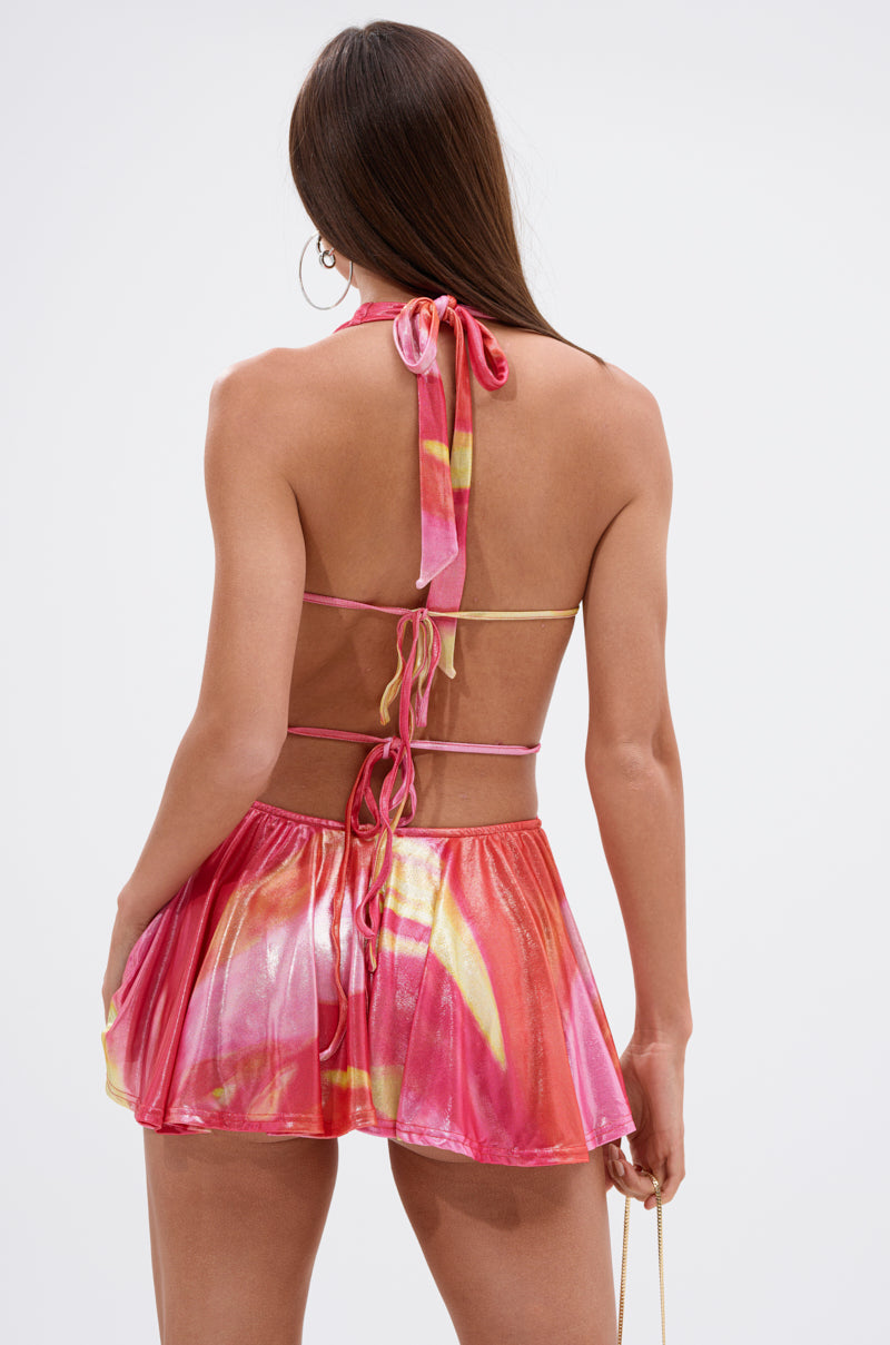 A woman with long brown hair is seen from behind wearing the YOU WISH PRINTED MINI DRESS, a vibrant abstract halter-neck festival mini dress with pink, yellow, and orange patterns, open back, and ties at the neck and lower back.