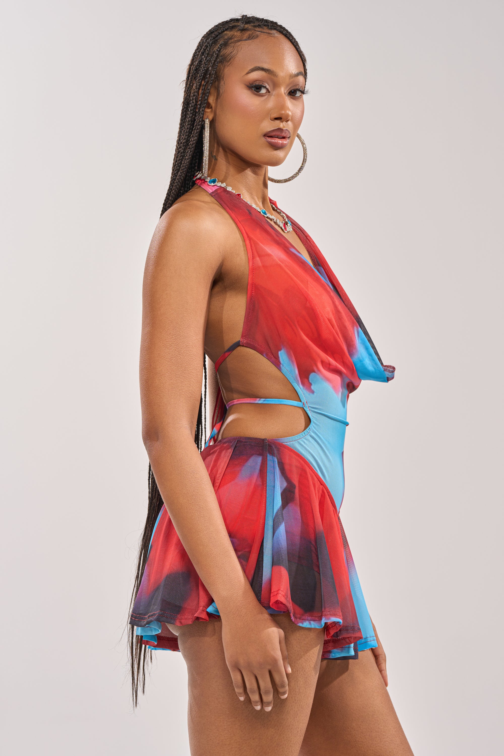 A woman models the BAMBI MINI SLEEVELESS DRESS, featuring an open back and red and blue waist cutouts. She wears long braids and large hoop earrings, posing against a plain light background. 5