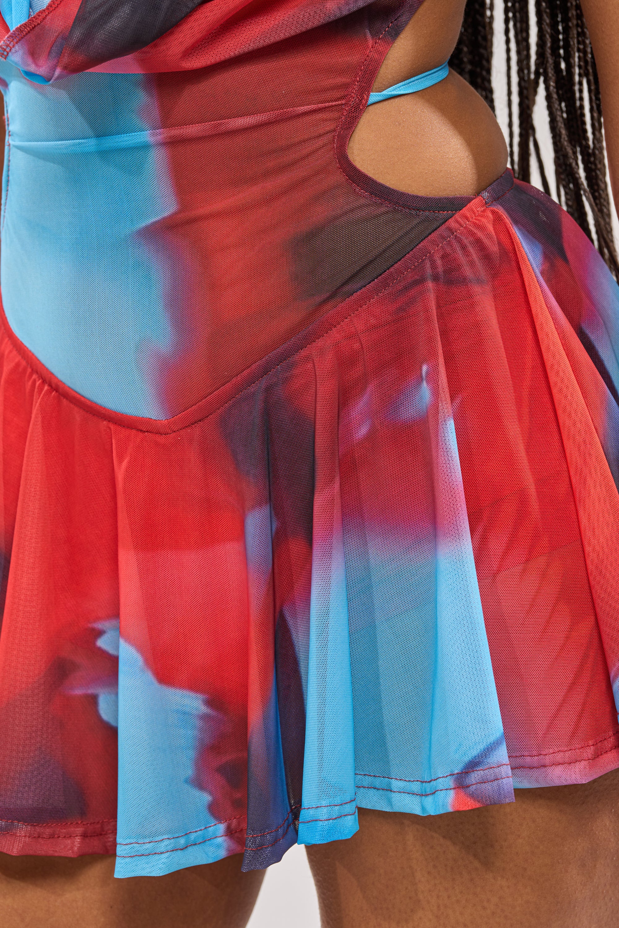 Close-up of a person with long braided hair wearing the BAMBI MINI SLEEVELESS DRESS, featuring an abstract watercolor print, mesh fabric, cut-out waist details, and a pleated mini skirt. 9