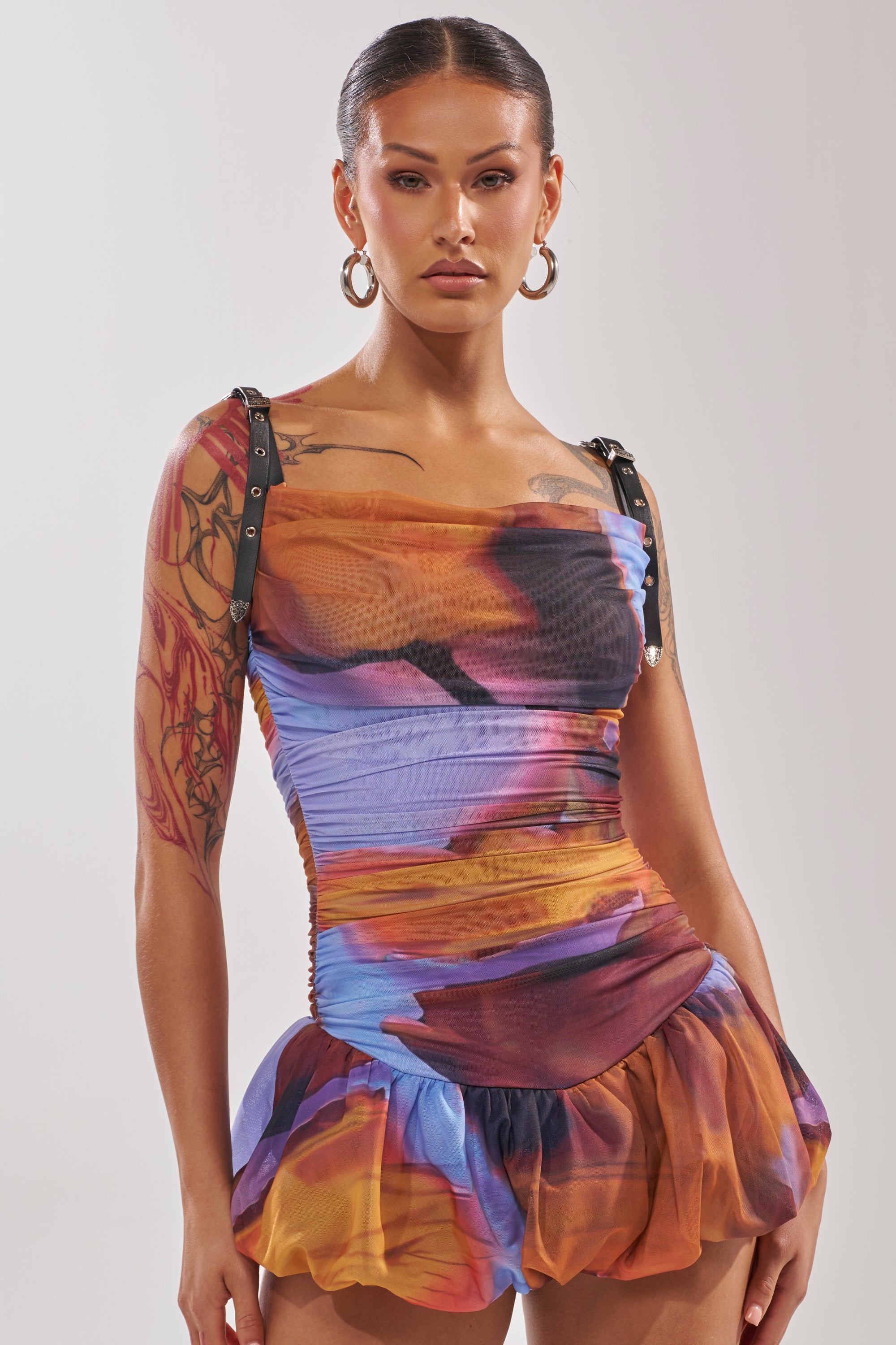 A person with hoop earrings and tattooed arms wears the IN A MOOD PRINTED MINI DRESS IN PURPLE—a colorful, form-fitting mini dress with black straps, cowl neck, ruffled hem, and bold blue, orange, and purple tie-dye print—against a light background. 0