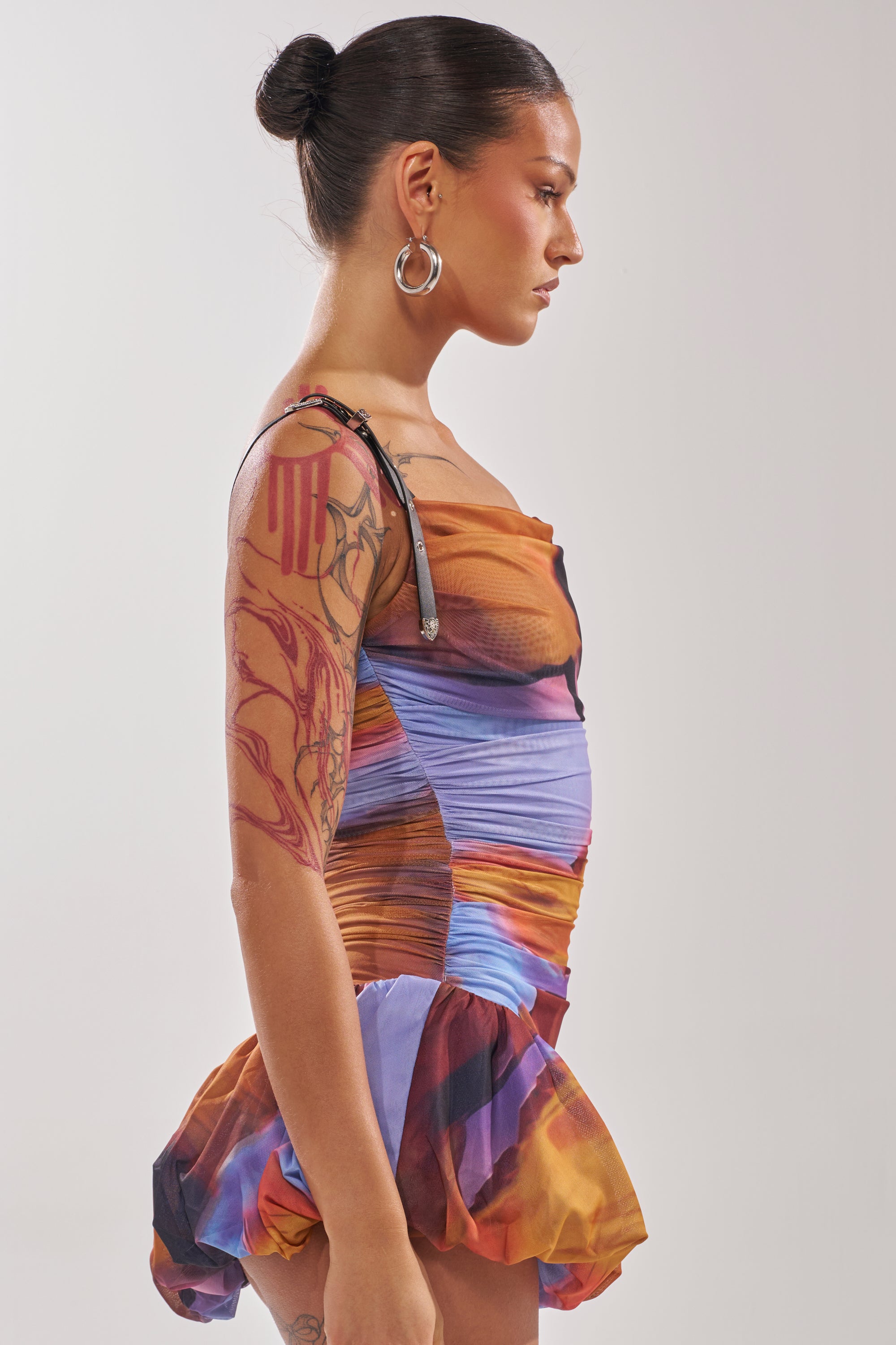 A woman with dark hair in a bun, large hoop earrings, and visible arm tattoos wears the IN A MOOD PRINTED MINI DRESS IN PURPLE. She stands in profile with a neutral expression against a plain background. 2