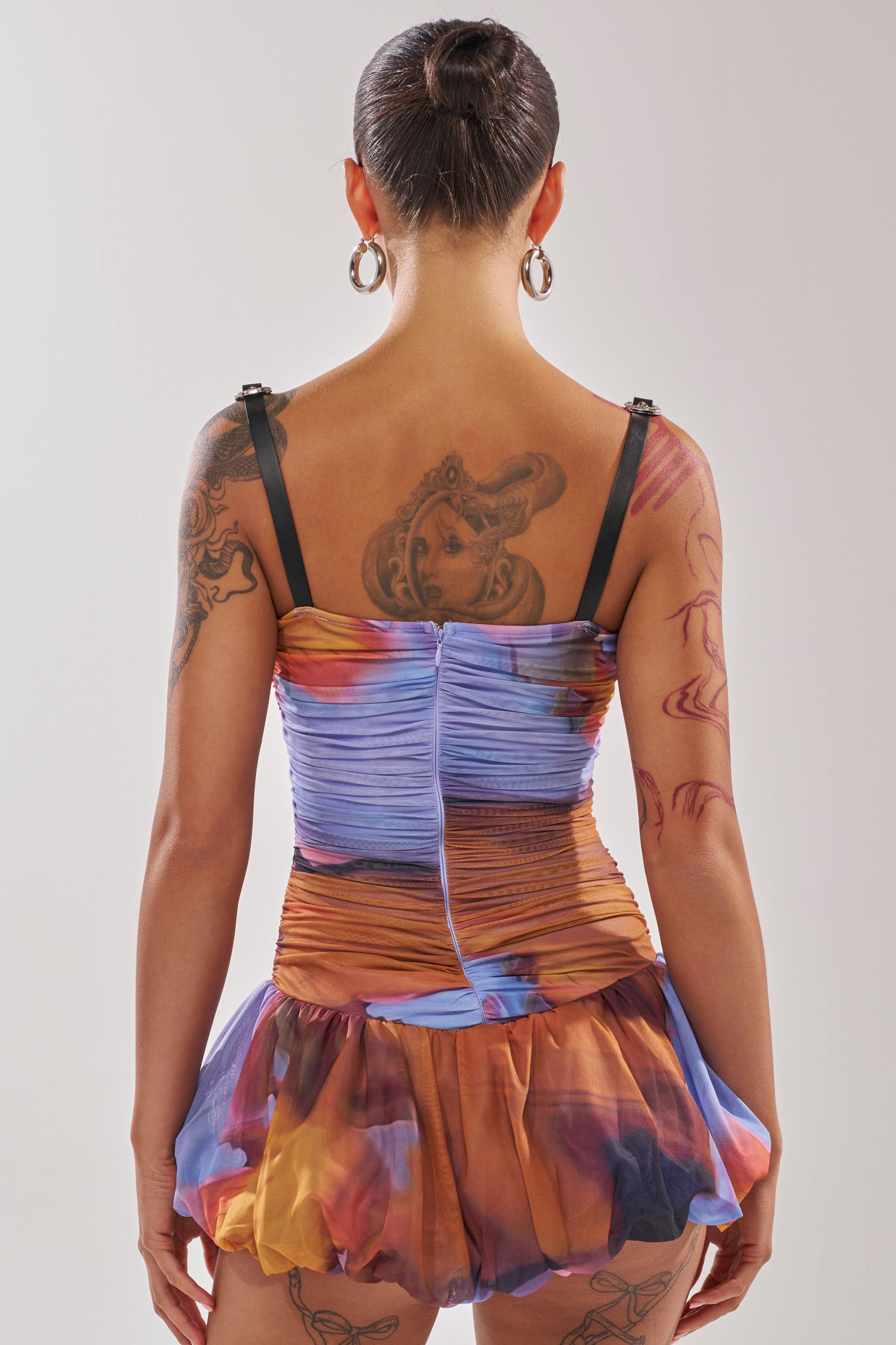 A person faces away, wearing the IN A MOOD PRINTED MINI DRESS IN PURPLE with thin black straps and a ruffled skirt. Their hair is in a bun, and they have large hoop earrings plus tattoos on their back and arms. 3