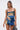 A woman with long straight brown hair models the IN A MOOD PRINTED MINI DRESS, featuring a square neckline, ruched details, and faux leather buckle straps, as she holds a black handbag against a plain white background.