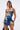 A woman with long straight hair poses against a white background wearing the IN A MOOD PRINTED MINI DRESS, featuring a blue, yellow, and black tie-dye pattern, faux leather buckle straps, and a ruffled hem.