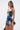 A woman with long straight hair in a high ponytail wears the IN A MOOD PRINTED MINI DRESS—a sleeveless, ruched mini with blue and beige florals and a flared ruffled hem—standing against a white backdrop holding a black handbag.