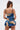 A woman with long brown hair in a high ponytail models the IN A MOOD PRINTED MINI DRESS, featuring blue and yellow ruffles and spaghetti straps, shown from the back against a plain white background.