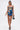 A woman wears the IN A MOOD PRINTED MINI DRESS featuring a blue and yellow abstract pattern, paired with a small black handbag and black heeled sandals, standing against a plain white background.