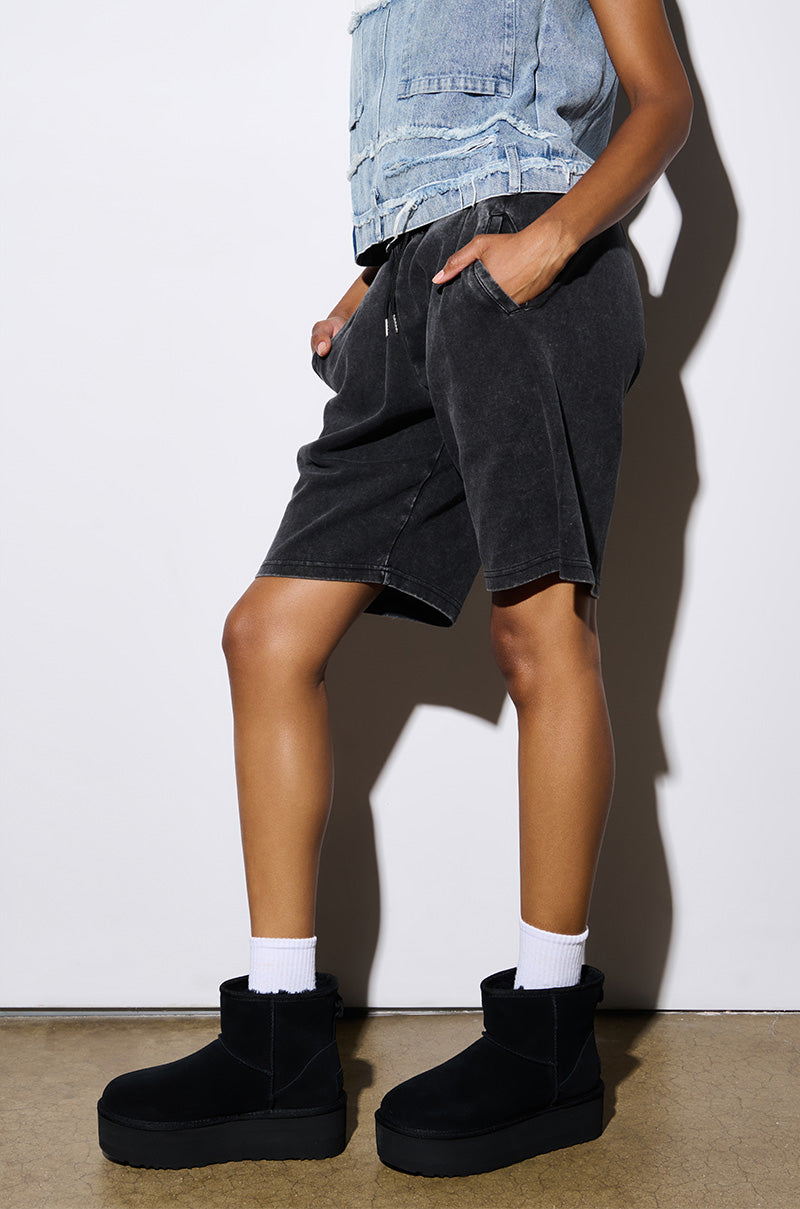 A person stands against a white background wearing a sleeveless denim top, dark shorts, and white socks. They are showcasing the fashionable UGG W Classic Mini Platform in Black with its distinctive platform sole. Their hand is tucked into one pocket, and their head is not visible in the image.