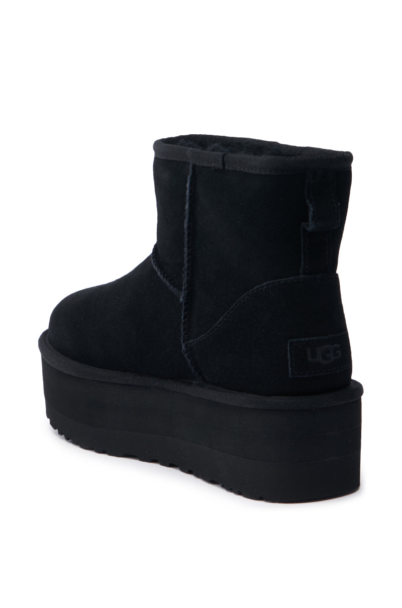 The UGG W CLASSIC MINI PLATFORM BLACK showcases a captivating black platform sole and plush inner lining, as seen from the rear and a subtly angled side view. Its wool blend upper is accentuated by a short shaft with prominent stitching, while the "UGG" logo is prominently featured on the heel.