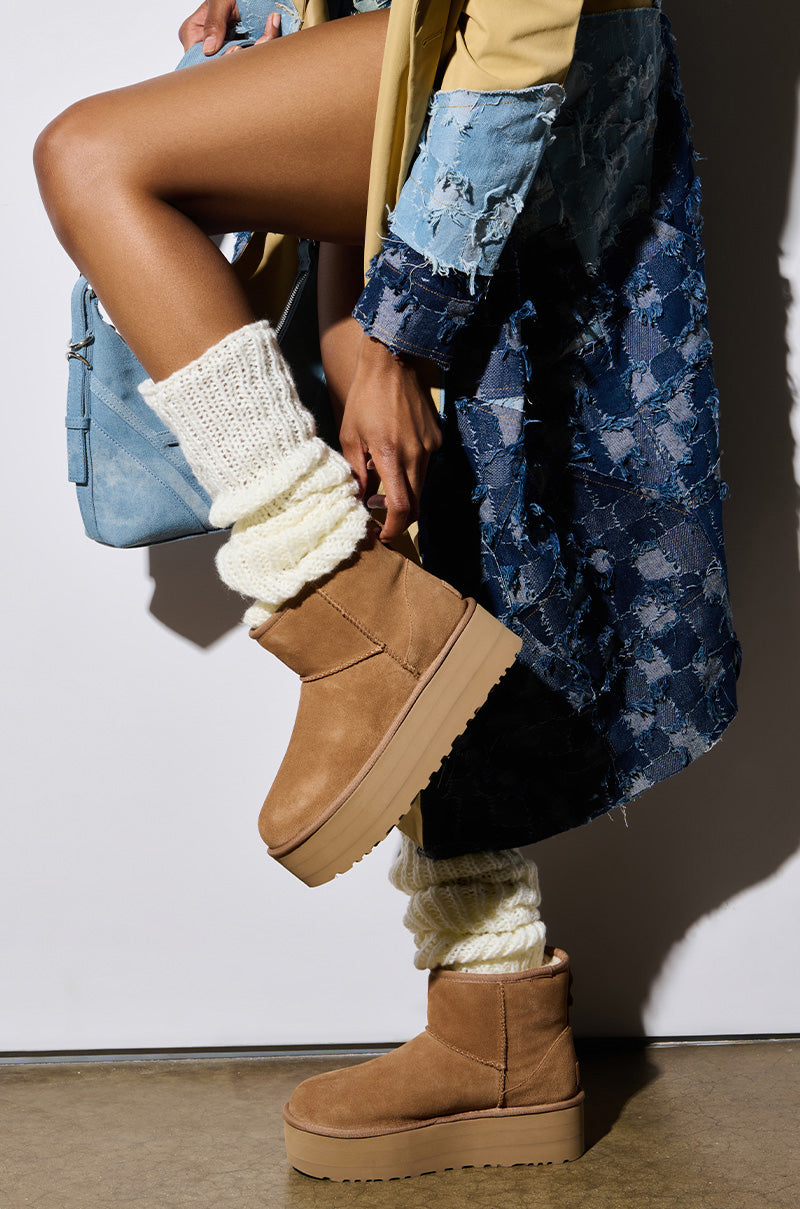 A person strikes a fashionable pose, highlighting the UGG Classic Mini Platform Bootie in Chestnut with its wool blend upper, paired perfectly with cream knit socks. They are dressed in a patterned blue coat and carry a light blue handbag, all set against a plain backdrop that enhances the ensemble's charm.