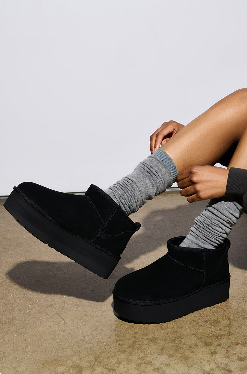 A person sits on a concrete surface against a white background, wearing the UGG Classic Ultra Mini Platform in black with a wool blend upper and gray slouchy socks. The chunky sole booties feature a simple design that contrasts elegantly with the textured socks.