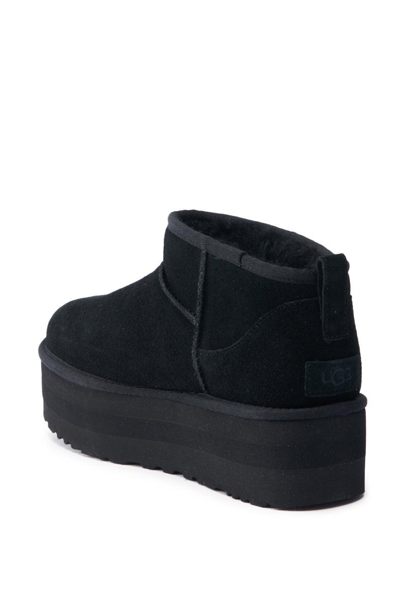 UGG CLASSIC ULTRA MINI PLATFORM IN BLACK features a black suede platform bootie with a sturdy sole, rounded toe, and subtle stitching details. The top edge showcases a wool blend upper for added warmth. The boot includes a pull tab at the back, reflecting a minimalist design reminiscent of the UGG Classic Ultra Mini.