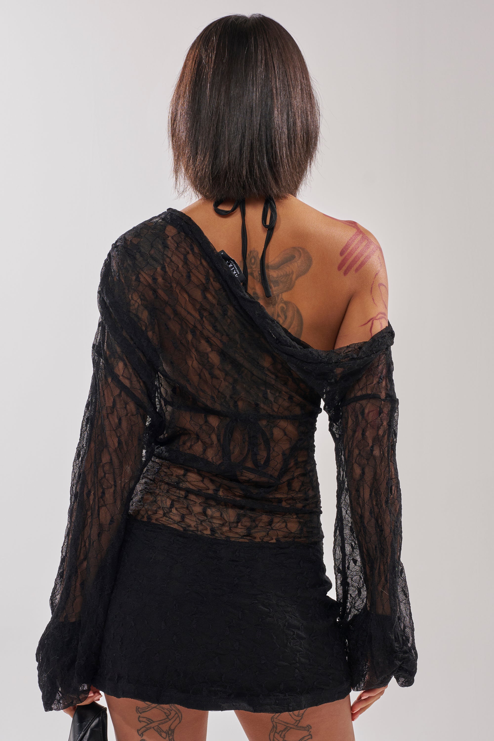A person with short dark hair, visible tattoos, and bare shoulders models the TO THE CLUB MINI DRESS with a black purse, standing back to the camera against a plain light background.
