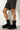 A person wearing black shorts and the chic UGG W CLASSIC DIPPER BLACK boots with yellow accents stands against a white brick wall. These boots boast a platform sole and a stylish design, ideal for making a bold statement.