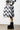 A person wearing a black and white wavy-patterned puffer coat and UGG BLACK W CLASSIC SWEATER LETTER boots stands in a minimalist indoor space, with their boots showcasing a cozy cable knit shaft and warm shearling lining as well as distinct yellow accents.