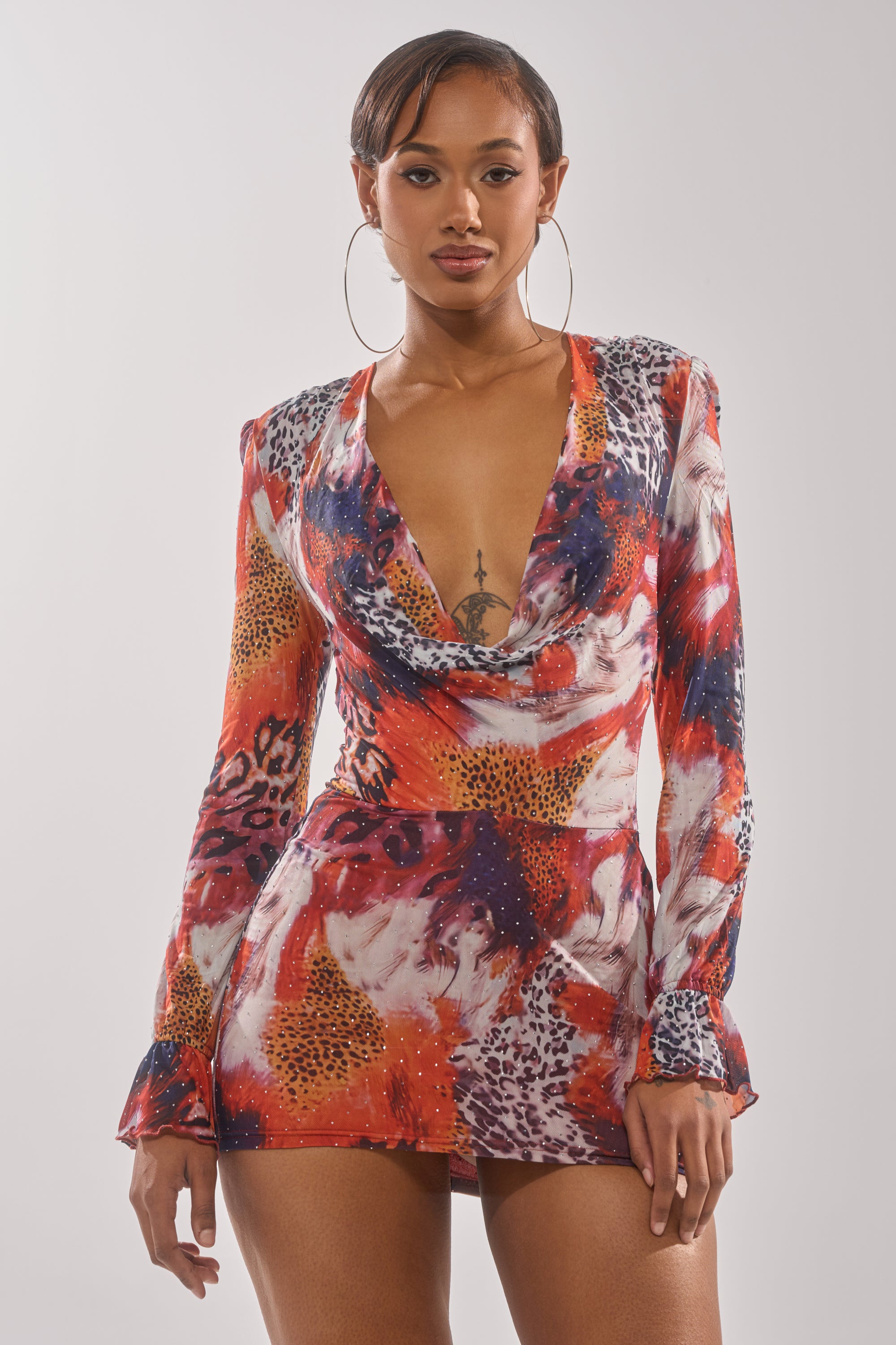 A woman poses confidently in the ON THE PROWL LEOPARD MINI DRESS—a vibrant bodycon mini with long sleeves, deep cowl neckline, and bold red, orange, and white leopard print—styled with hoop earrings and a chest tattoo. 0