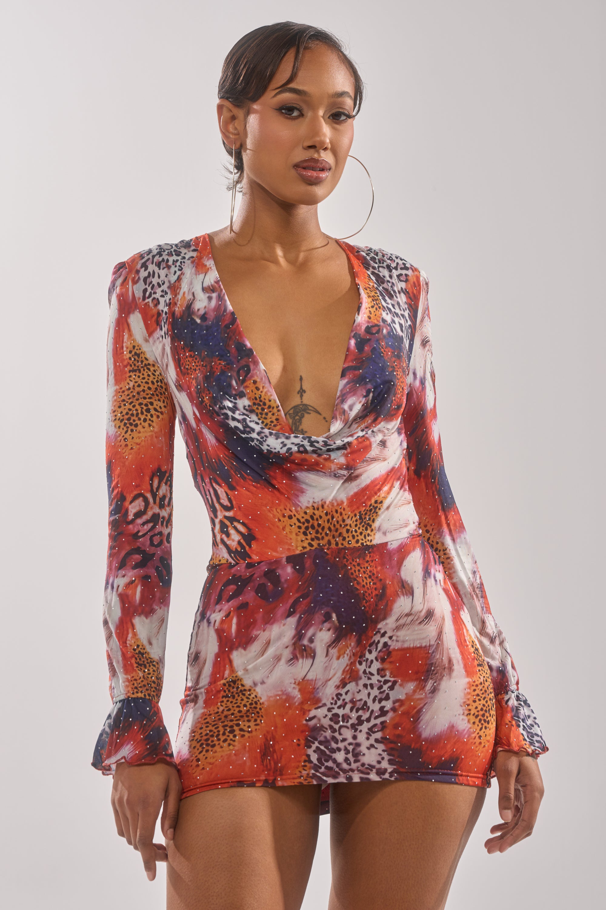 A woman confidently poses against a plain light background, wearing the ON THE PROWL LEOPARD MINI DRESS, featuring a vibrant leopard print, long sleeves, and a deep cowl neckline. 1