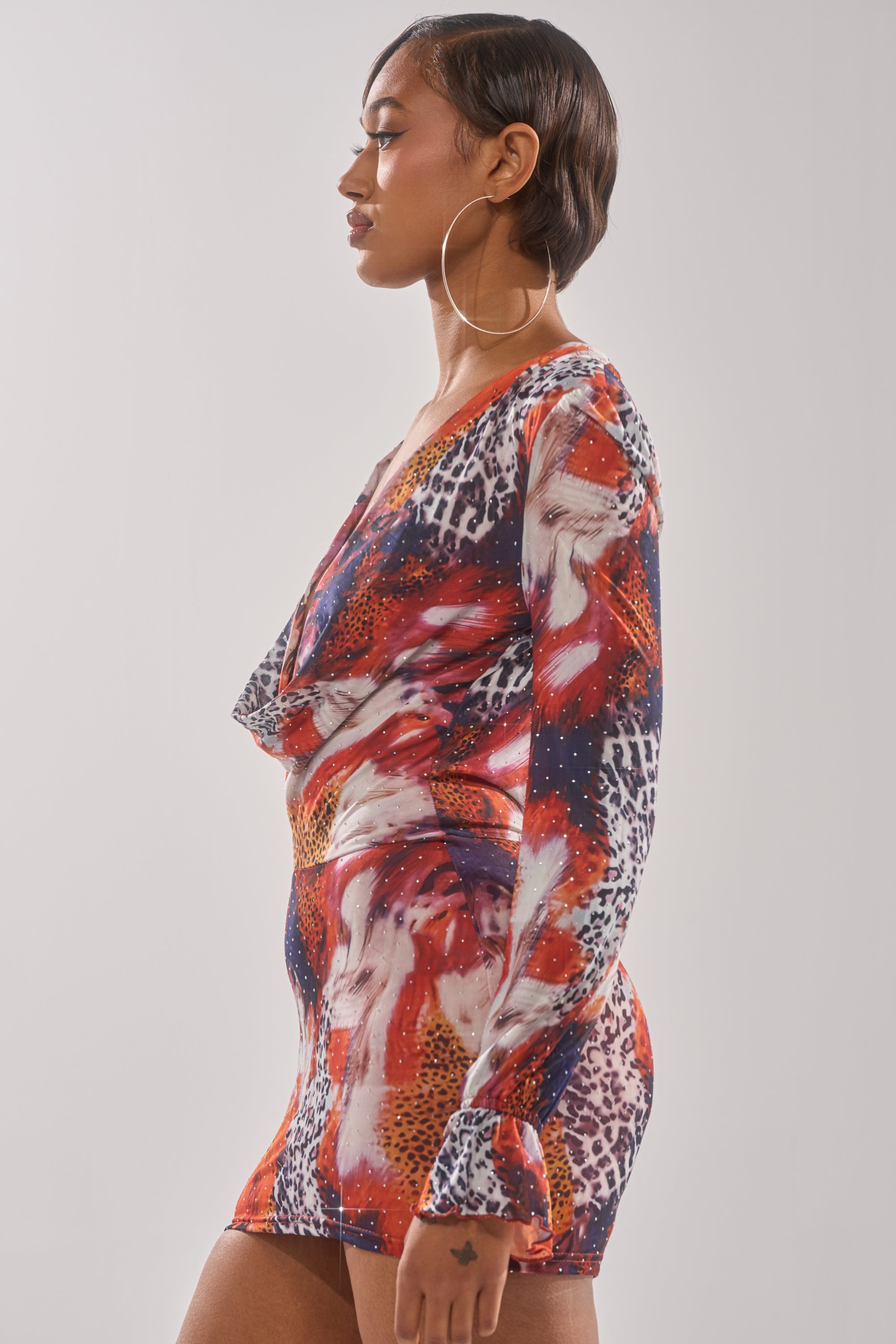 Woman in profile wears the ON THE PROWL LEOPARD MINI DRESS, a long-sleeve, multicolored animal print style with a draped neckline and large hoop earrings, standing against a plain light background. 3