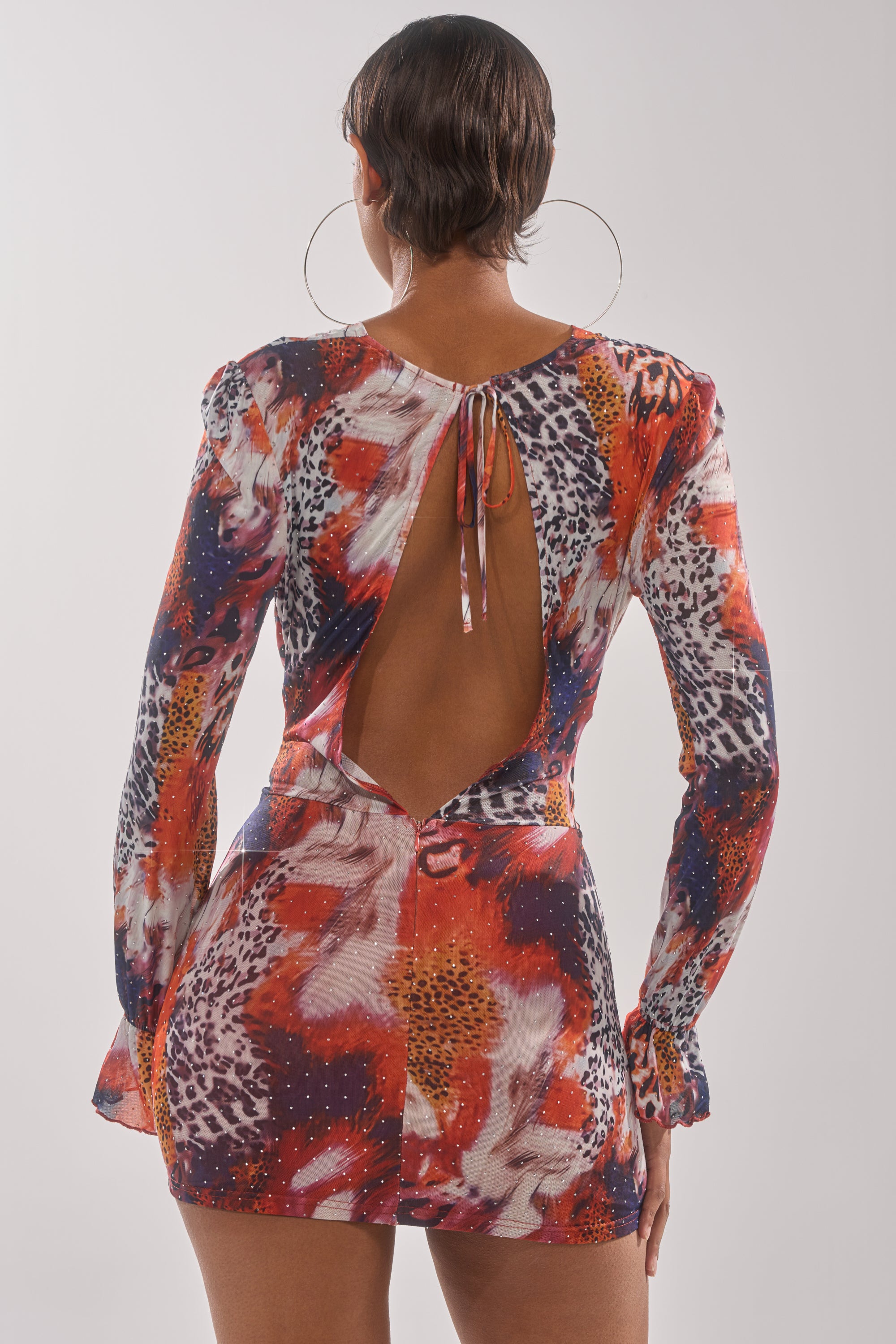 A person with short hair and hoop earrings wears the ON THE PROWL LEOPARD MINI DRESS, featuring a colorful bold animal print, while standing with their back to the camera against a plain light background. 4
