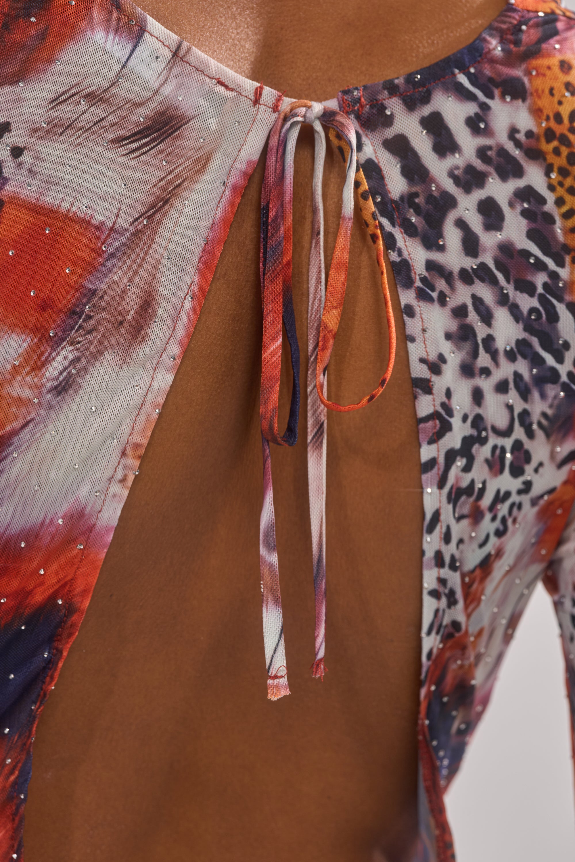 Close-up of a person wearing the ON THE PROWL LEOPARD MINI DRESS, featuring a colorful open-back design with an upper back tie and an abstract red, orange, black, and white leopard print accented by sparkling details. 7