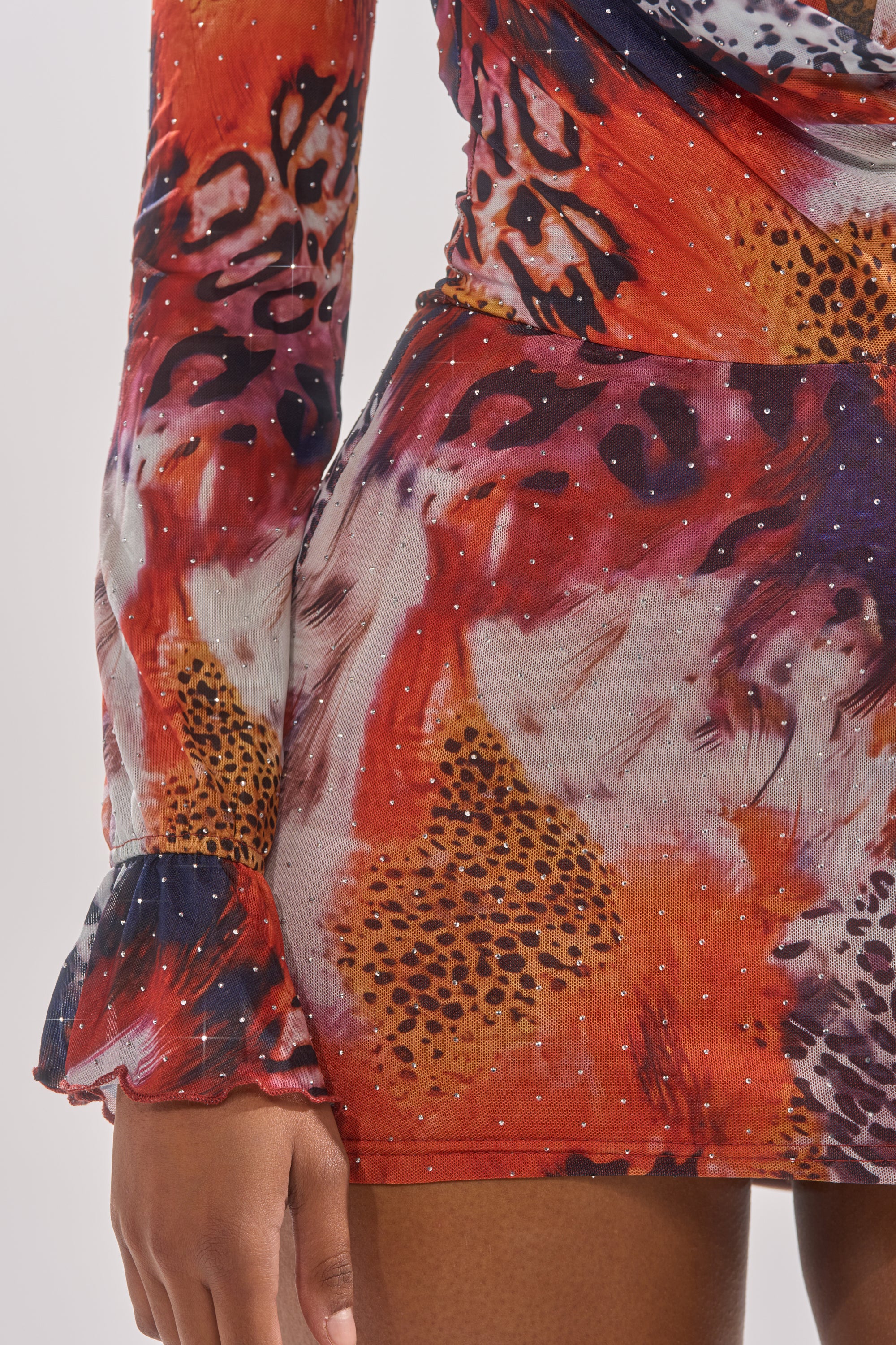 A close-up of a person wearing the ON THE PROWL LEOPARD MINI DRESS, featuring vibrant red, orange, and purple hues with sparkling details, highlighting the lower arm, hand, and part of the skirt. 8
